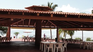 An outdoor patio with tables and chairs, a covered area with a tiled roof, and a view of the sea.