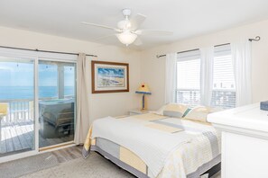 A bedroom with a bed, a ceiling fan, a window with curtains, a picture on the wall, and a view of the ocean through a sliding door.