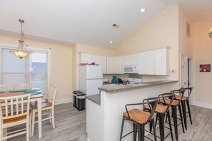 Cocina compacta con gabinetes blancos, microondas y encimera de granito. Hay una zona de comedor con mesa y sillas, y un espacio con taburetes que tiene cuatro taburetes.
