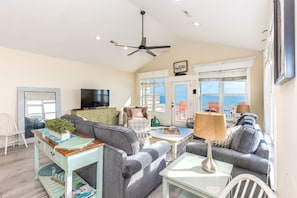 A cozy living room with a ceiling fan, a television, and a view of the sea through large windows.
