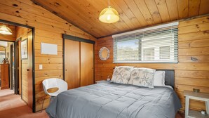 A wooden cabin bedroom with a bed, a chair, a bedside table, and a window with blinds.