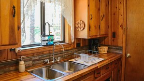 A wooden kitchen with a double sink, a window with lace curtains, and a wooden cabinet with a decorative plate. - Family Cabin in the Pines. All Wood! Charming memory-maker. (Pinetop-Lakeside)