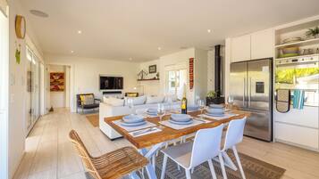 A modern kitchen with a stainless steel refrigerator, a dining area with a wooden table and white chairs, and a living area with a sofa and a TV.