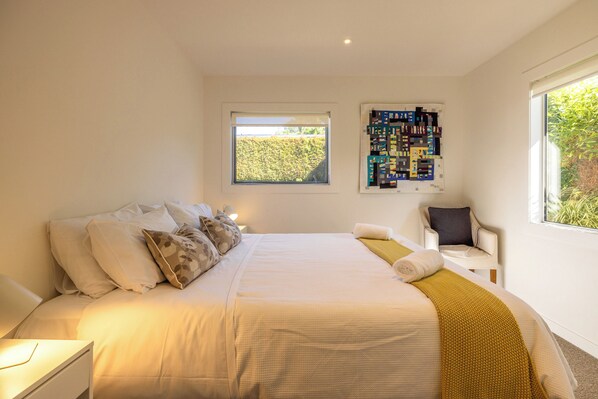 A bedroom with a large bed, a nightstand, a chair, a window with a view of greenery, and a wall-mounted board with various items. - Moa Magic - Oneroa Holiday Home (Oneroa)
