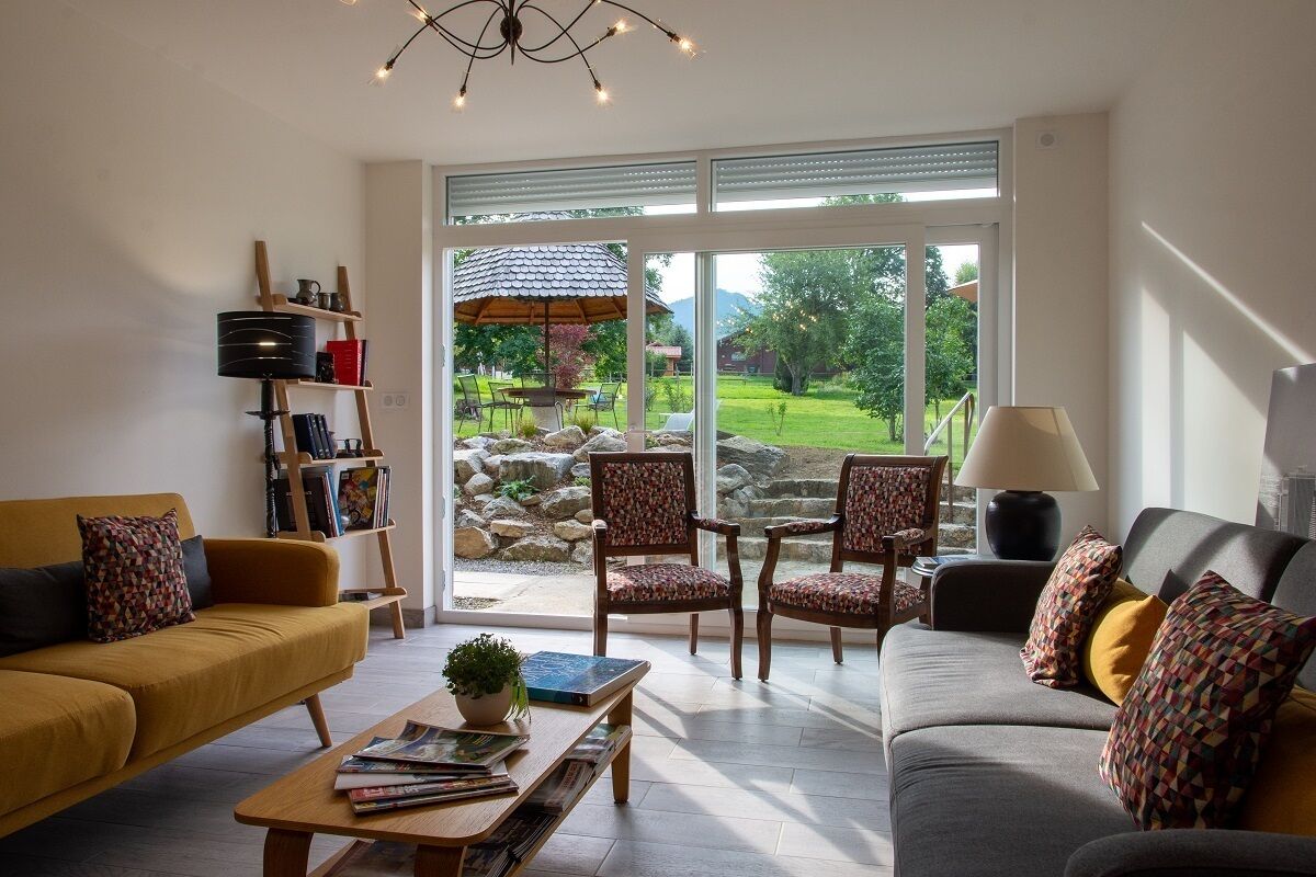 A living room with two sofas, a coffee table, and a bookshelf. There is a large window with a view of a garden and a patio.