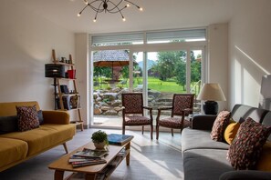 A living room with two sofas, a coffee table, and a bookshelf. There is a large window with a view of a garden and a patio.