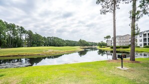 A serene riverside view with a grassy area, a small water body, and a building with multiple floors in the background. - Sandpiper 2A Sunset Beach (Sunset Beach)