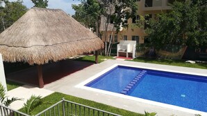 Una piscina con una struttura a tetto di paglia e lettini, circondata da vegetazione e un edificio sullo sfondo.