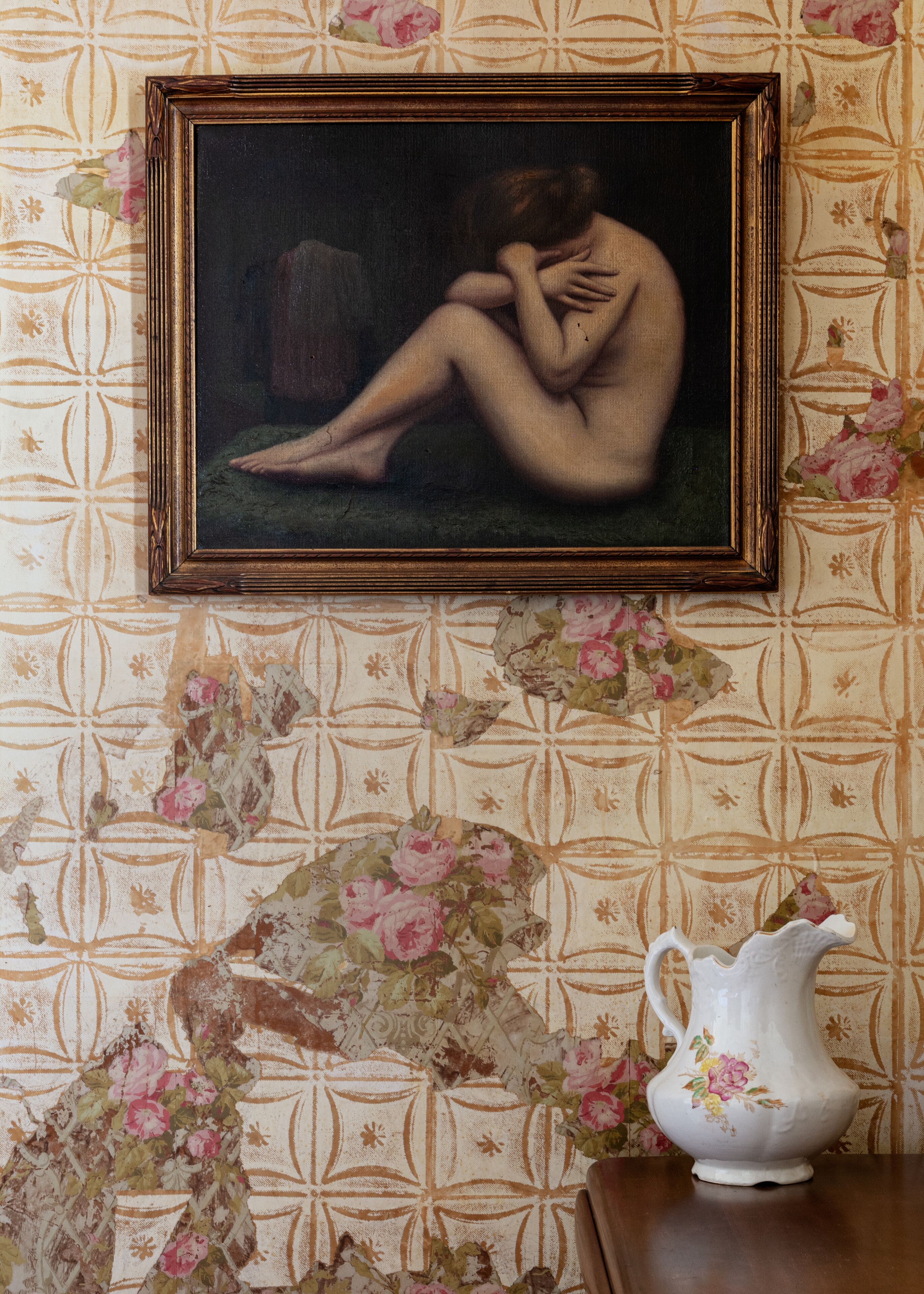 A framed painting of a nude figure on a wall, a floral-patterned wallpaper, and a white pitcher with floral designs on a table.