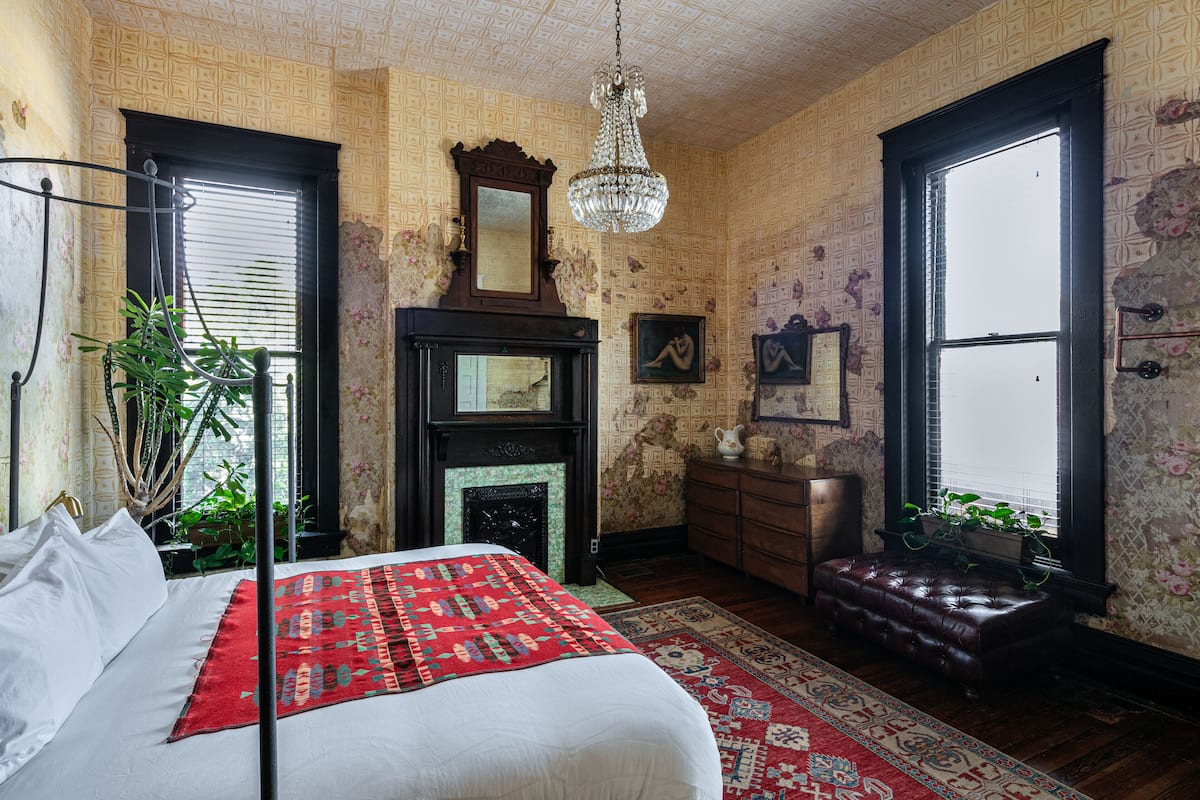 A bedroom with a bed, a fireplace, a chandelier, and a large window.