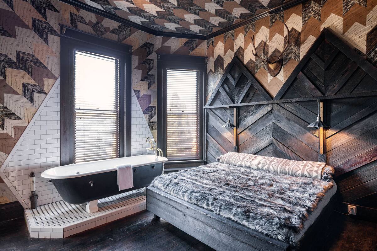 A modern bathroom with a freestanding bathtub, a large bed with a fluffy blanket, and a geometric-patterned wall.
