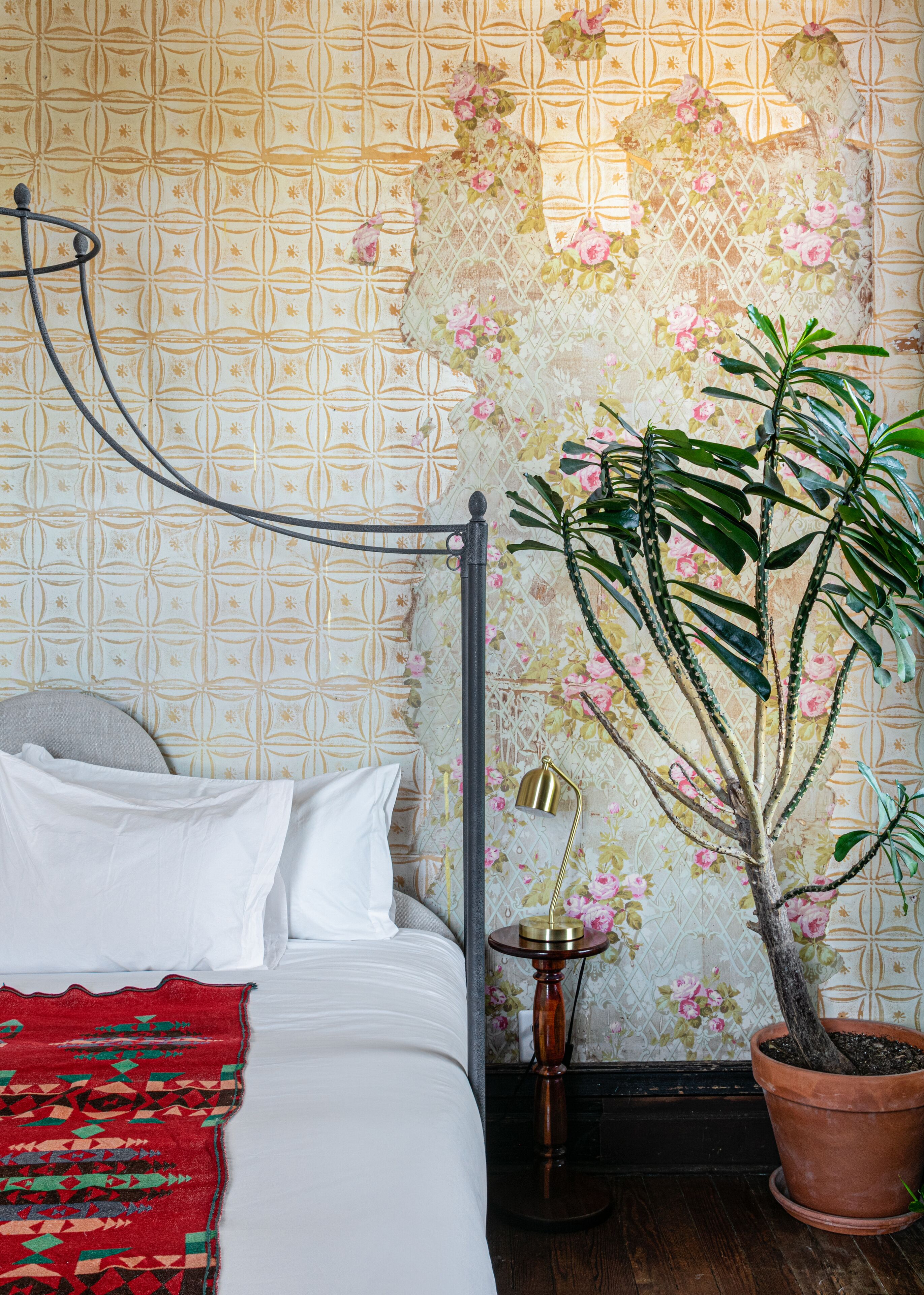 Un letto con lenzuola bianche, una coperta rossa con motivo decorativo e un poggiatesta in metallo. C'è una pianta in vaso con foglie verdi e fiori rosa.