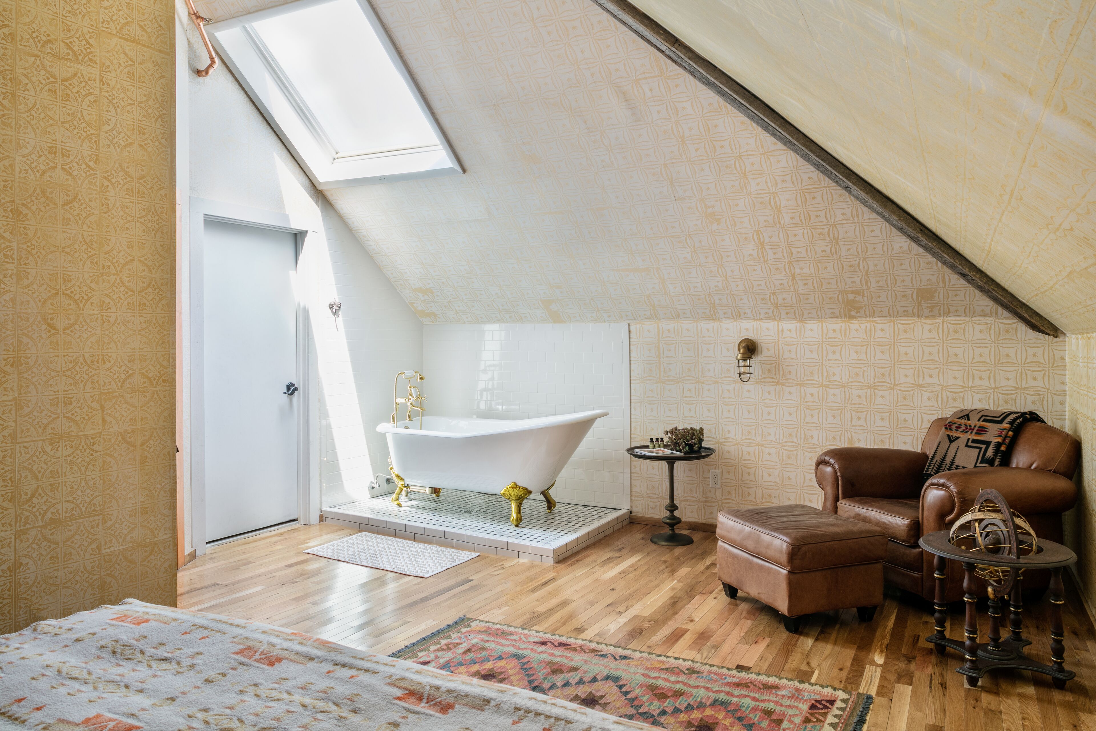 A room with a sloped ceiling, a white bathtub, a brown leather sofa, and a patterned rug.