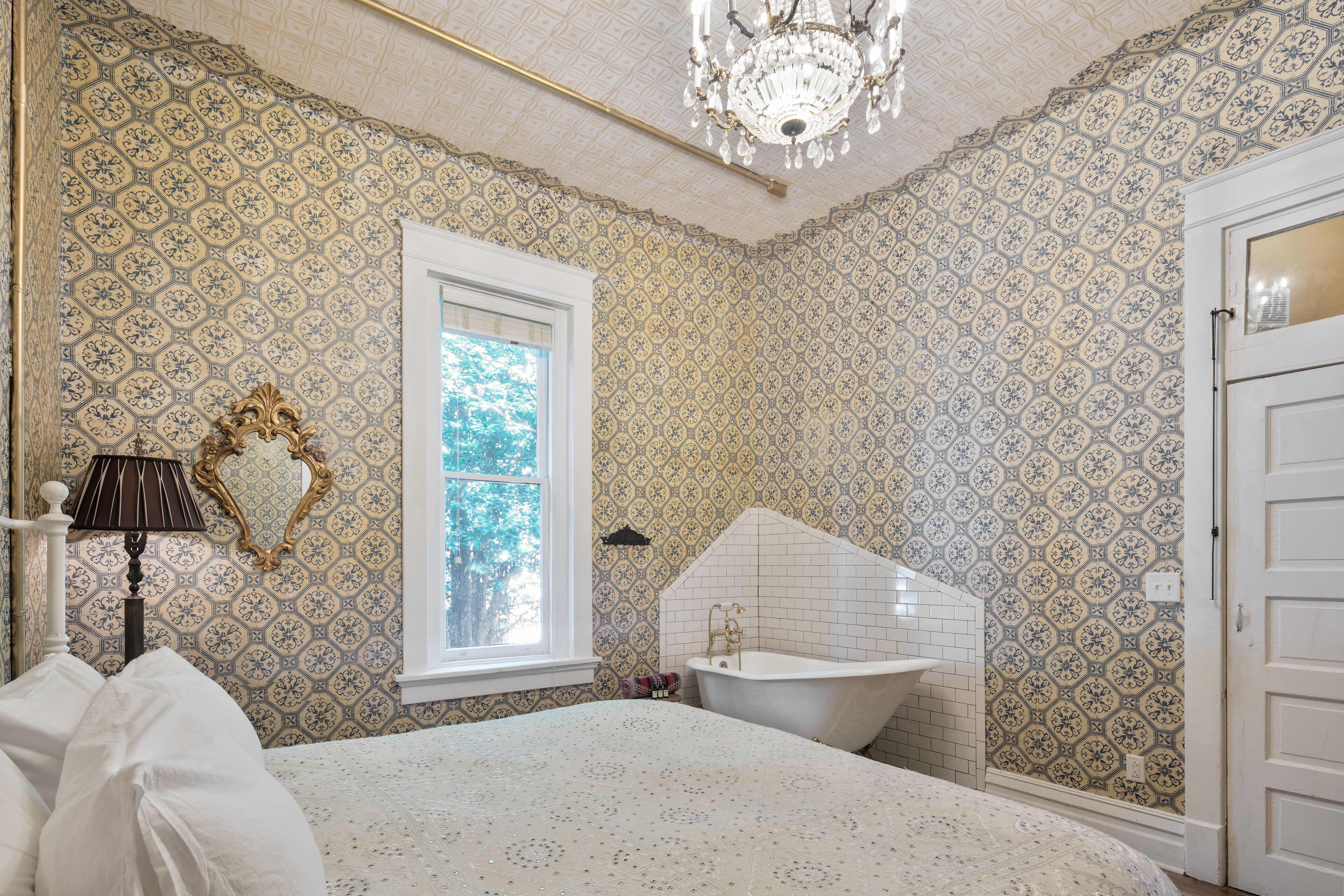 A bathroom with patterned wallpaper, a freestanding bathtub, a chandelier, and a window with a view of trees.