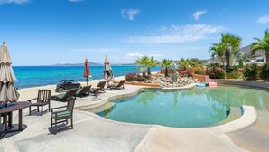 A pool area with lounge chairs and umbrellas, a clear view of the ocean, and a rocky landscape with palm trees.