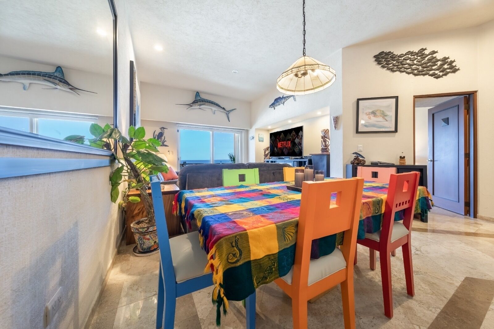 A dining area with a colorful quilted tablecloth, a potted plant, and a view of the ocean through a balcony railing.