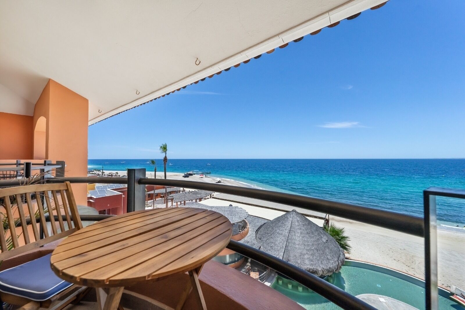 A balcony with a wooden table and chairs, overlooking a beach with clear blue water and a sandy shore.