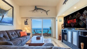 A living room with a leather couch, a coffee table, and a TV on a cabinet. There is a large shark decoration on the wall and a view of the ocean through the sliding glass door.