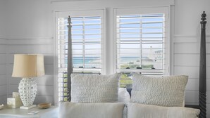 A bedroom with a bed, two windows, a nightstand, a lamp, and a view of the sea. - Buckeye on the Beach | Gulf Front | Seaside, FL | Right next to Town Center, Sleeps 10! (Santa Rosa Beach)