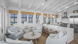 A modern kitchen with white cabinetry, a central island, and a dining area with white furniture. - Buckeye on the Beach | Gulf Front | Seaside, FL | Right next to Town Center, Sleeps 10! (Santa Rosa Beach)