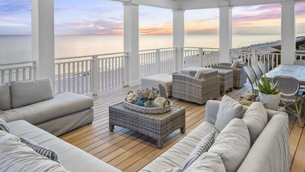A covered outdoor seating area with wicker furniture, a wooden deck, and a view of the ocean at sunset. - Buckeye on the Beach | Gulf Front | Seaside, FL | Right next to Town Center, Sleeps 10! (Santa Rosa Beach)