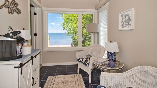 A cozy room with a view of the sea, a wicker chair, a small table with a lamp, and a kitchen area with a kettle and mugs.