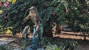 Un jardin agrémenté d’une sculpture représentant un poisson et un oiseau posés sur un rocher, le tout entouré de fleurs roses et de verdure.