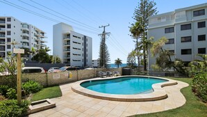 A swimming pool with a curved edge, surrounded by a brick wall and a grassy area. There are lounge chairs and a small deck area.