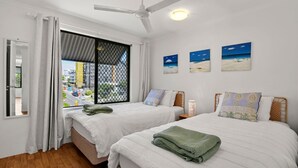A hotel room with two beds, a large window with a view of a cityscape, a ceiling fan, and three paintings on the wall.