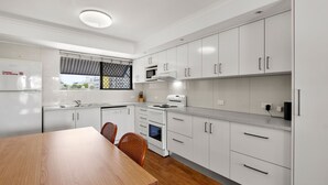 A modern kitchen with white cabinets, a stove, microwave, and a dishwasher. There is a dining table with chairs and a window with a view of buildings.