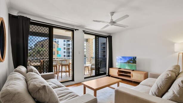 A modern living room with a sofa, a coffee table, and a TV on a wooden cabinet. There is a sliding glass door leading to an outdoor balcony with chairs and a table.