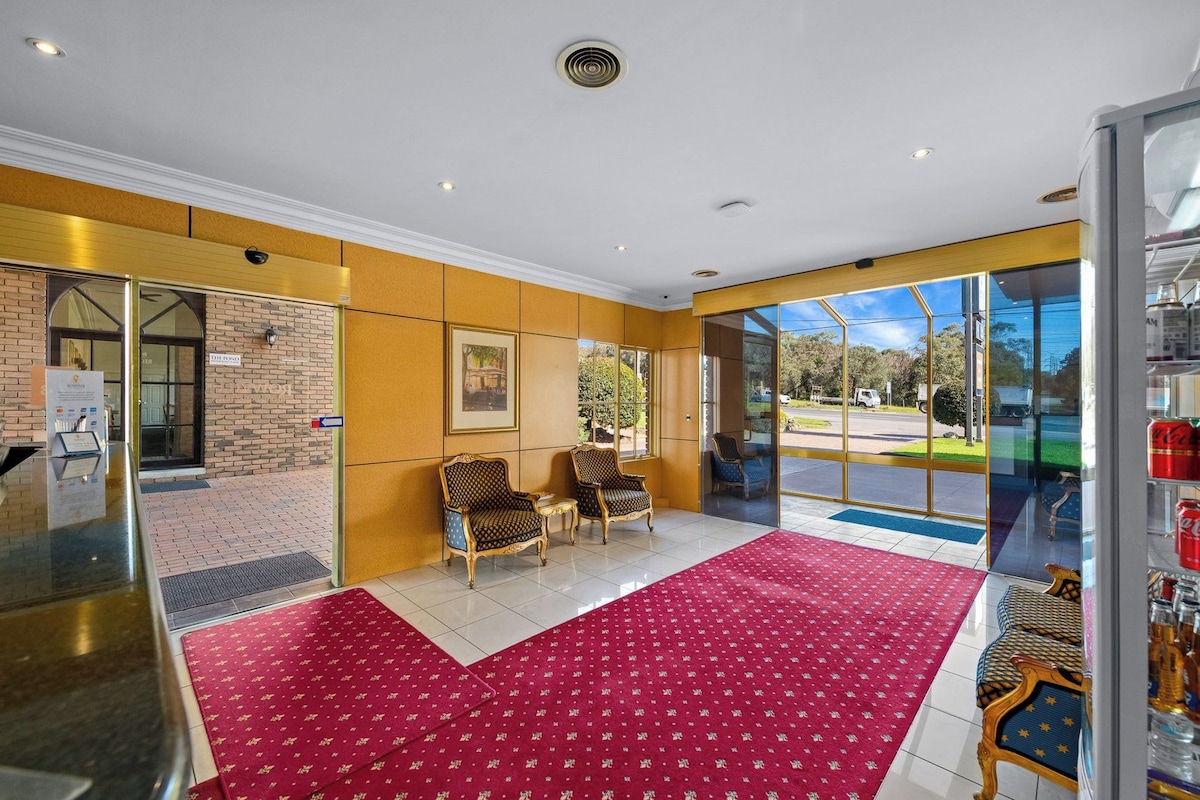 A hotel lobby with a red carpet, two armchairs, a reception desk, and a glass door leading outside.