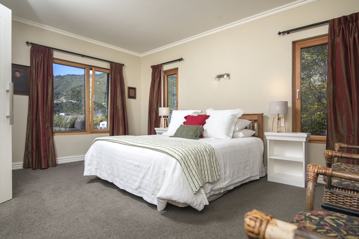 A bedroom with a large bed, two bedside tables, a nightstand, and a view of a mountainous landscape through the windows.