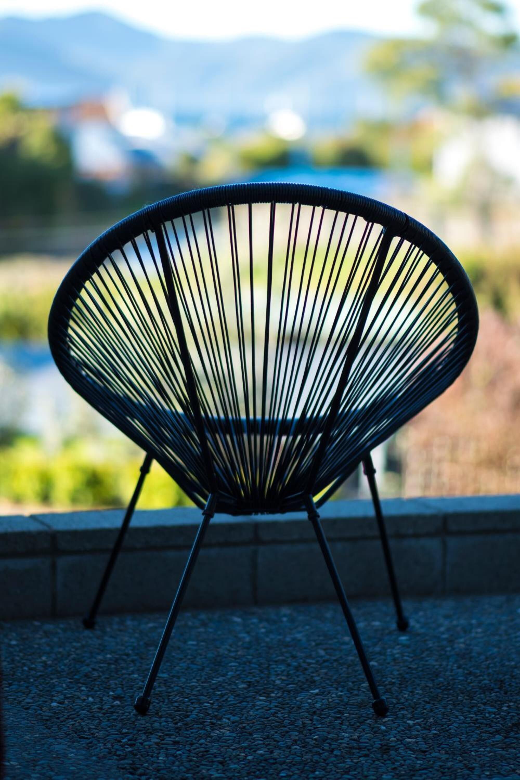 Una silla con un diseño de estructura metálica distintiva, ubicada al aire libre con un fondo desenfocado de follaje y un cuerpo de agua.