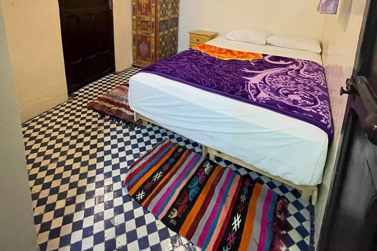 A bedroom with a checkered floor, a bed with a purple and white bedspread, a decorative ceiling light, and a wardrobe with intricate patterns.