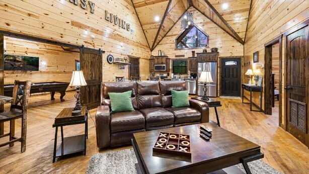 A spacious living area with a high ceiling, wooden walls, a leather sofa, and a coffee table with a game. - Easy Living -Mountain Setting w/ Pool Table & Private Hot Tub🎱🌲♨️ (Broken Bow)