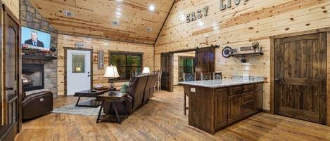 A cozy cabin interior with a fireplace, a television, a dining area, and a kitchen with wooden cabinets and a bar.