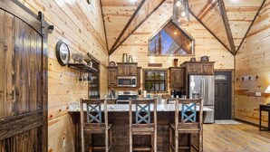 A rustic kitchen with wooden walls, a central island with bar stools, and a large window above the sink. - Easy Living -Mountain Setting w/ Pool Table & Private Hot Tub🎱🌲♨️ (Broken Bow)