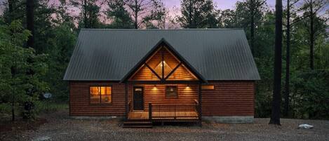 A log cabin with a gabled roof, a front porch, and a gravel driveway.