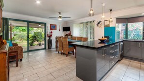 A modern kitchen with a central island, dining area with wicker chairs, and a large sliding glass door leading to a garden. - Delightful Duplex on Rose (BONGAREE)