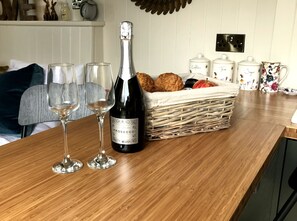 Una mesa de madera con una botella de Prosecco, dos copas, una cesta de pan y utensilios de cocina.