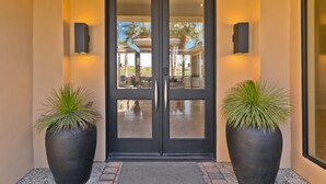 Une porte double noire avec des panneaux de verre, flanquée de deux grands pots de plantes, et un petit espace extérieur aménagé en coin salon, visible à travers la porte.