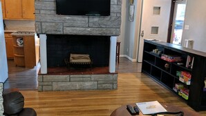 A living room with a stone fireplace, a flat-screen TV mounted above it, a brown leather couch, a wooden coffee table, and a bookshelf filled with books and games. - CHARMING NEIGHBORHOOD IN PURDUE'S BACKYARD (West Lafayette)