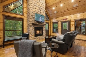L’intérieur chaleureux d’un chalet en bois comprend une cheminée, des fauteuils en cuir et de grandes fenêtres qui offrent une vue sur la forêt environnante.