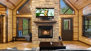 Interior acolhedor de uma cabana com lareira de pedra, paredes de madeira, uma TV grande fixada acima e uma área de estar confortável.