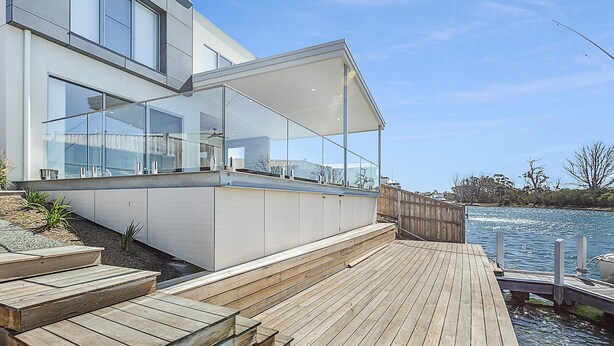 A modern house with a glass balcony overlooking a body of water, a wooden deck, and a clear sky.