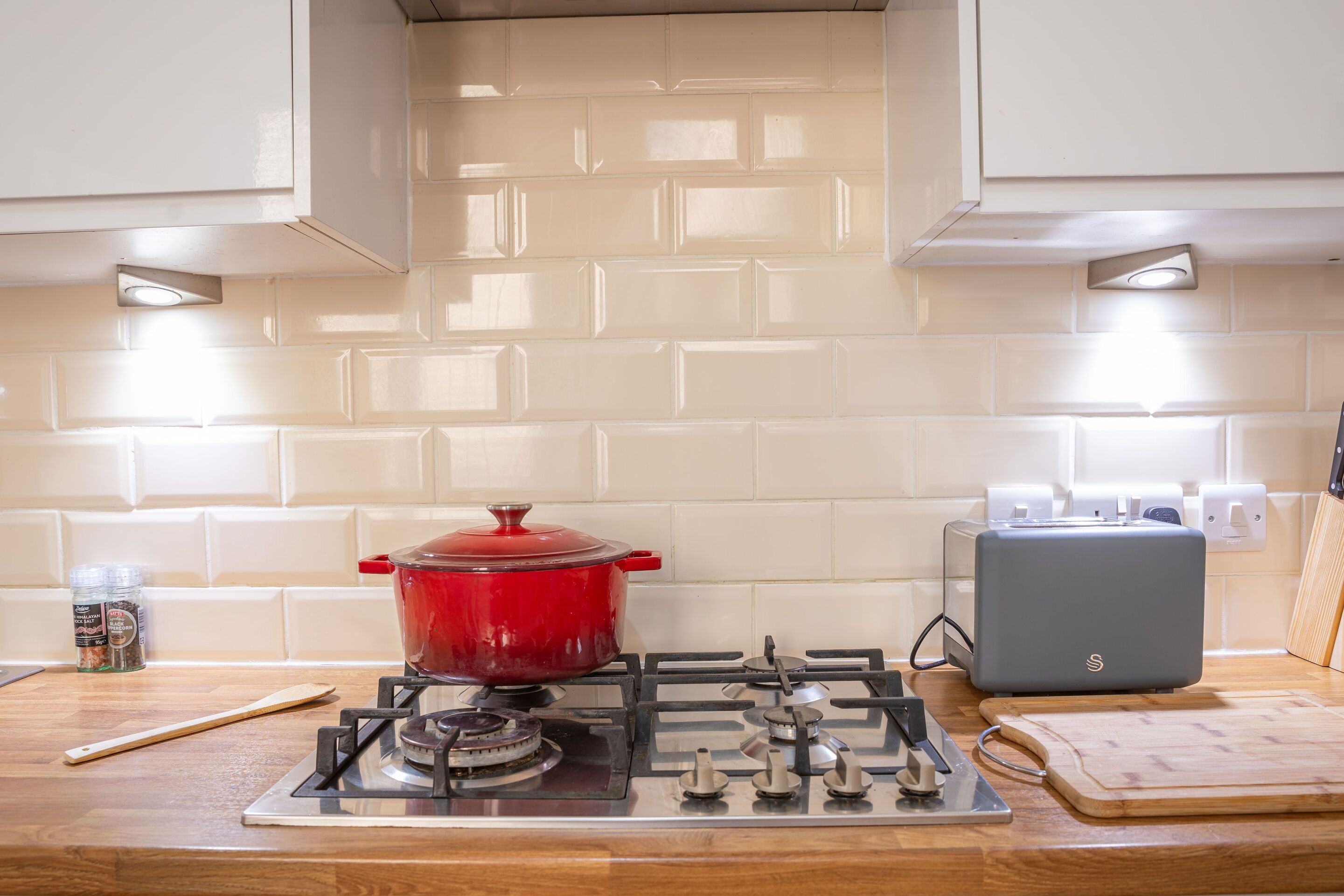 Een keuken met een gasfornuis, een rode pan, een broodrooster en een houten snijplank.