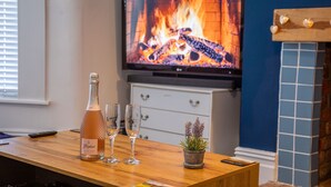 A wooden bar area with a bottle of wine, two glasses, and a small potted plant. A television displaying a fireplace is mounted on the wall.