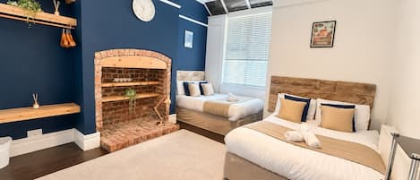 A bedroom with a brick fireplace, two beds, a large window with blinds, and a clock on the wall.