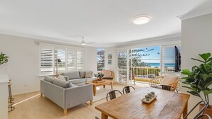 A spacious living area with a grey sofa, a wooden dining table, and a balcony with a view of the sea. - Beach Pines 5 - Fully Renovated North Kirra Beachfront - Min. 3 Night Stays (Bilinga)